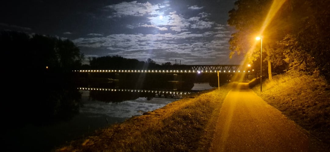 Noční pohled na asfaltovou stezku podél řeky osvětlenou oranžovou pouliční lampou. Vlevo se v klidné vodě zrcadlí osvětlený železný most a nad ním svítí měsíc mezi mraky.