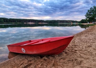 Červená záchranářská loďka se znakem červeného kříže leží na písečném břehu jezera. V klidné vodě se zrcadlí zatažená obloha a za jezerem se táhne borový les.